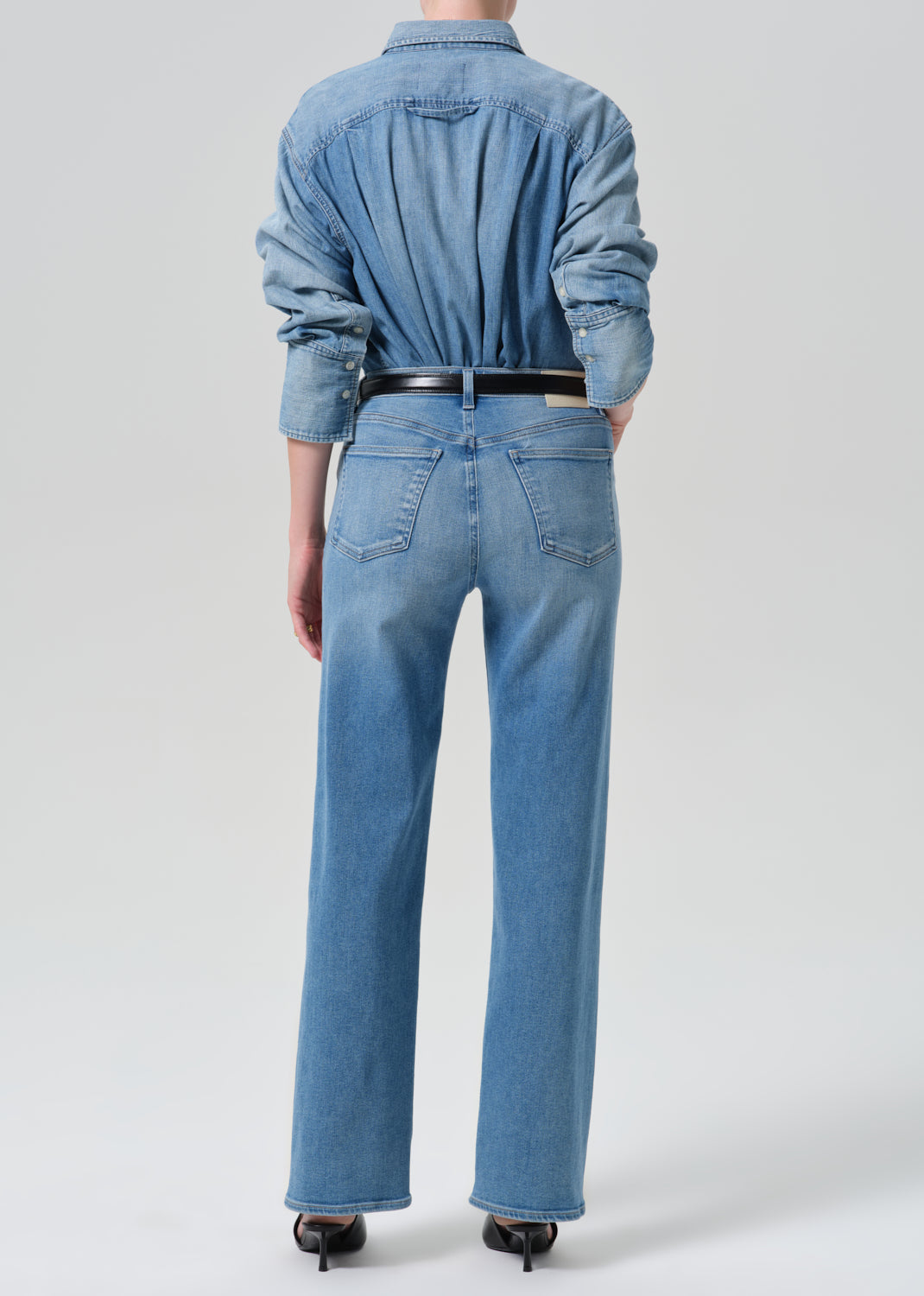 Person wearing a light blue denim shirt with light blue straight leg jeans viewed from the back on a grey background