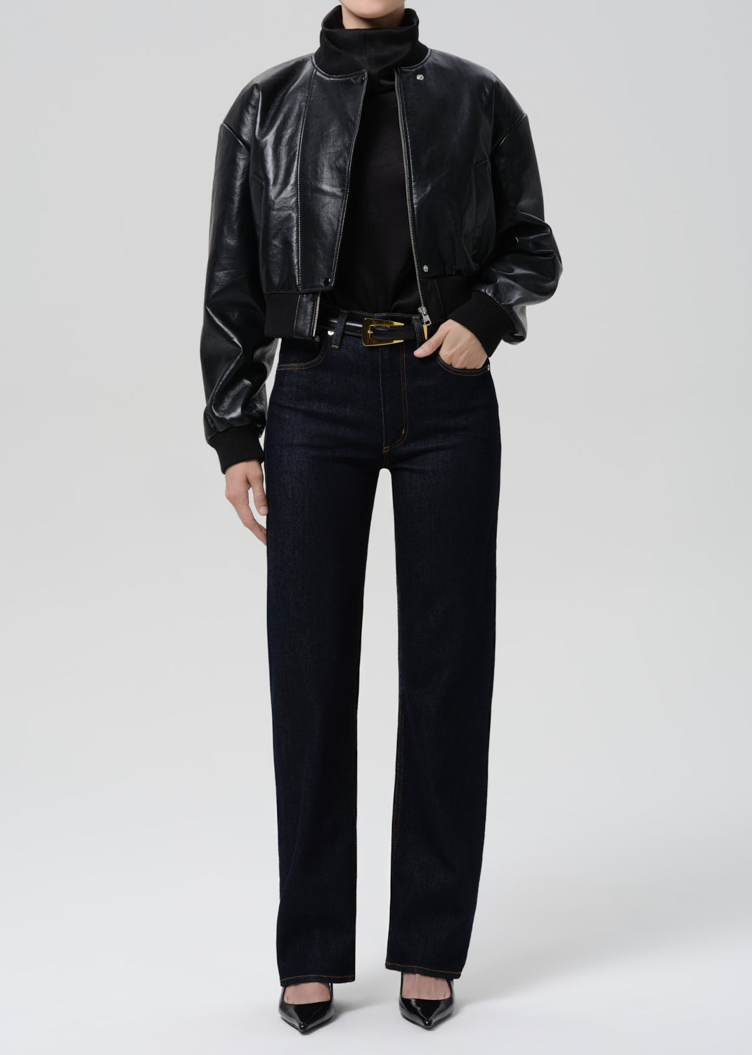 Person wearing a black leather jacket, black turtleneck, black jeans, and black shoes on a grey background