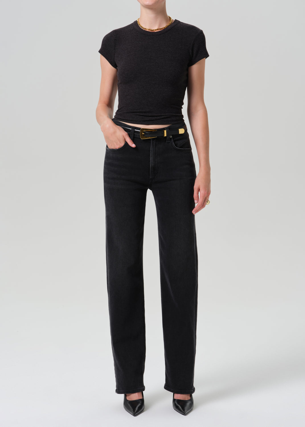 A model wearing a black short-sleeve crop top and black wide-leg jeans with a black belt, standing against a grey background.