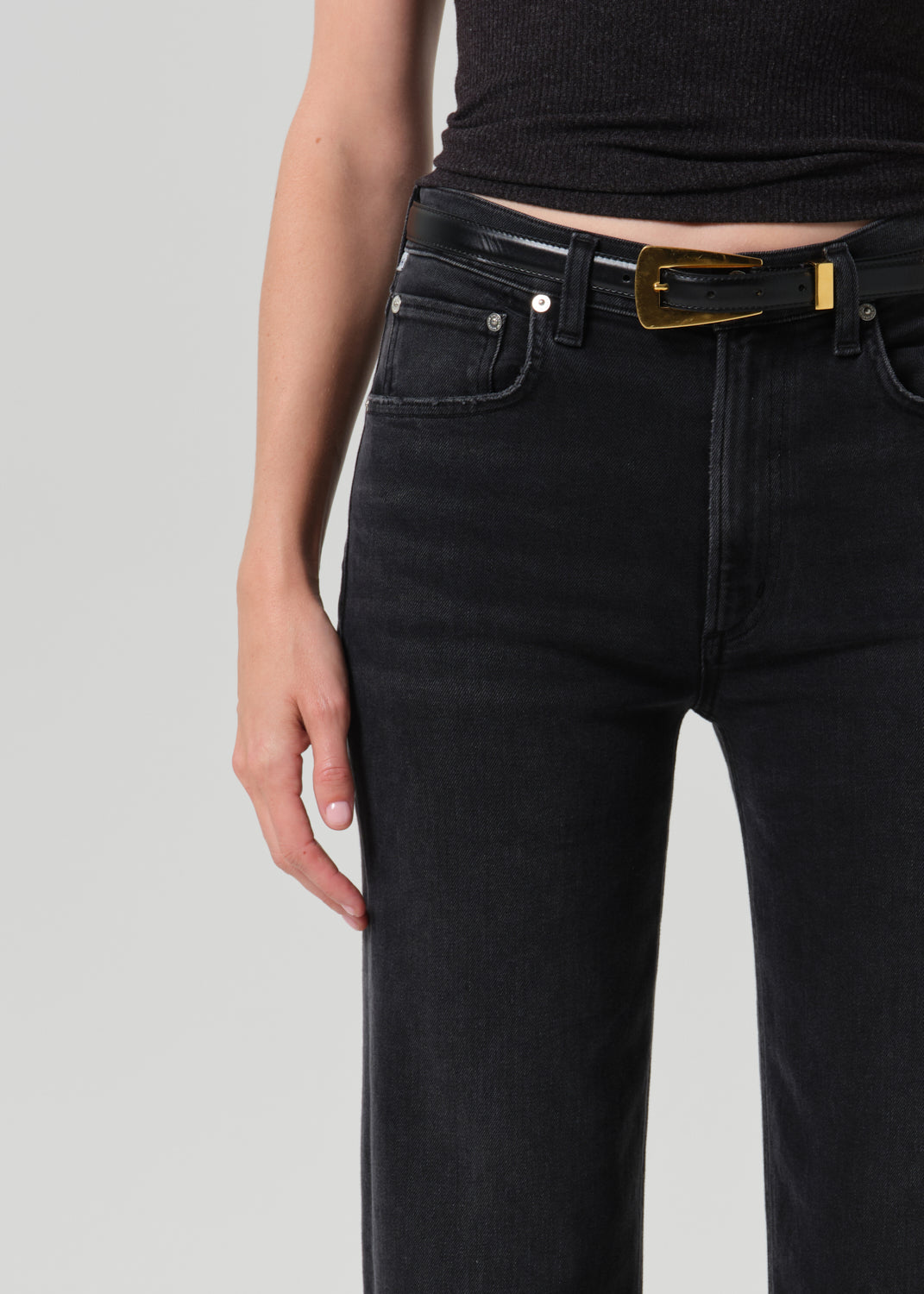 Close-up of a person wearing black jeans with a black belt featuring a prominent gold rectangular buckle, paired with a black ribbed top against a grey background.