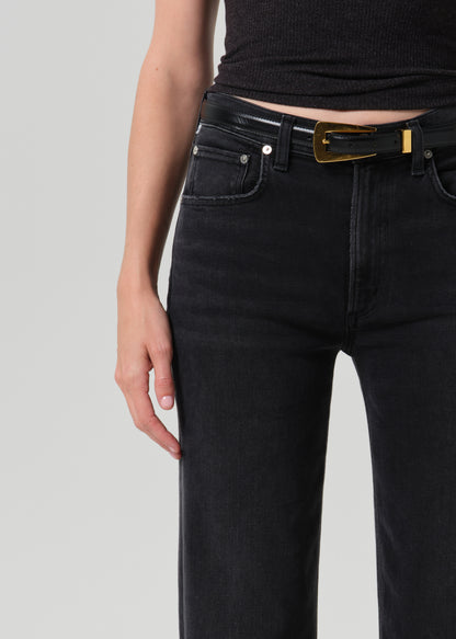 Close-up of a person wearing black jeans with a black belt featuring a prominent gold rectangular buckle, paired with a black ribbed top against a grey background.