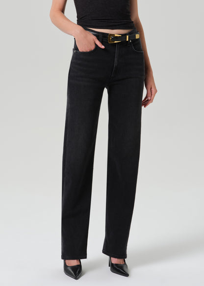 A model wearing black wide-leg jeans with a gold-tone belt, paired with a black crop top and black pointed-toe heels, shown from the waist down on a grey background.