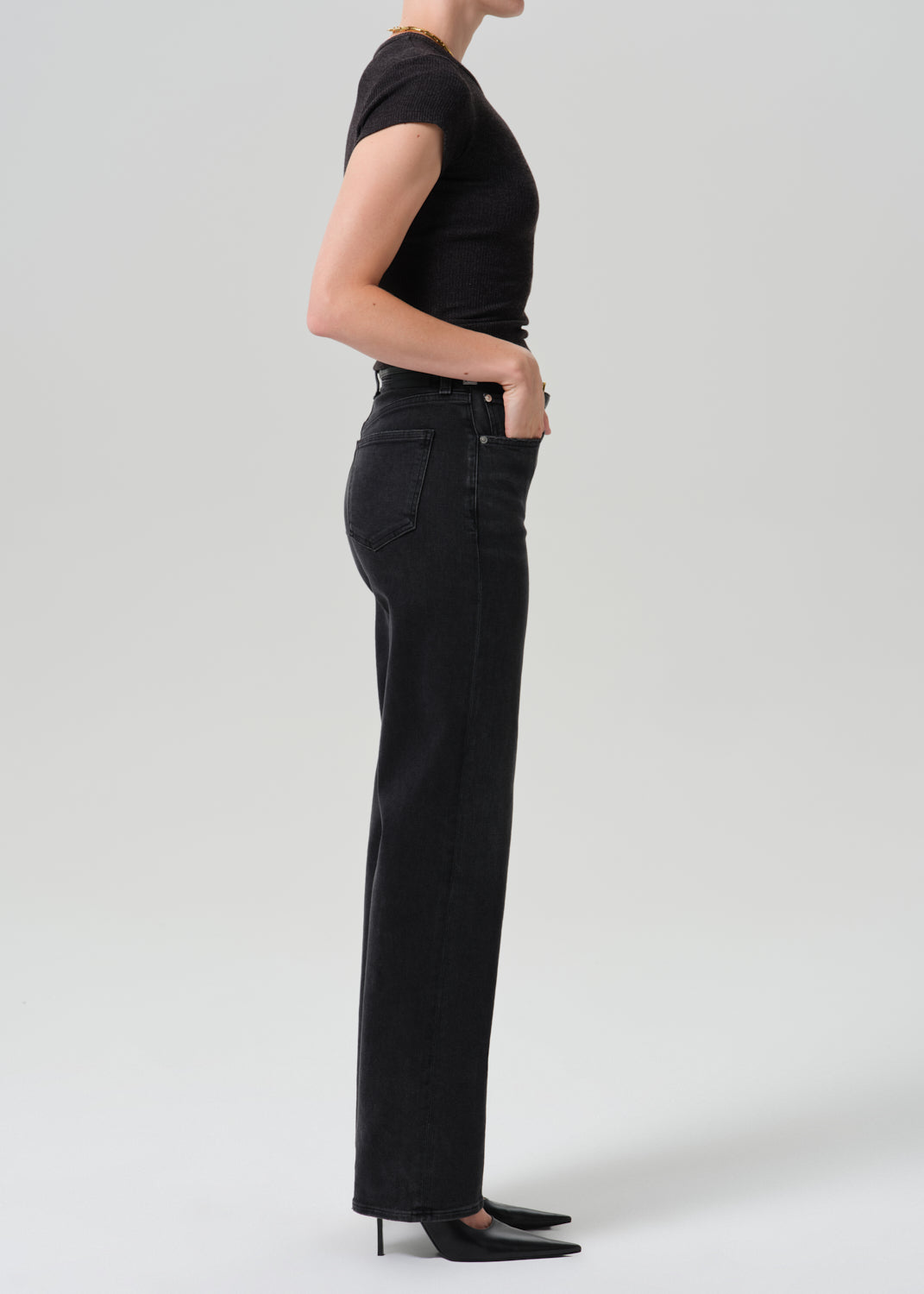 A side-profile studio shot of a model wearing black wide-leg jeans, a black short-sleeve top, and black pointed-toe heels against a grey background.