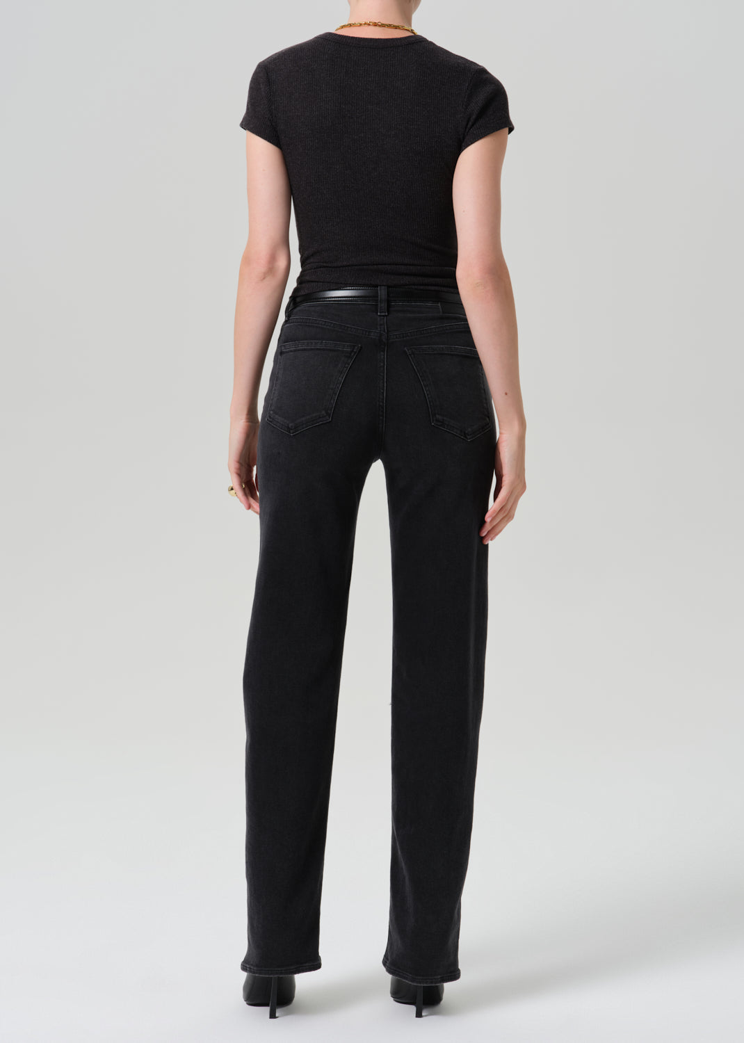 Rear view of a model wearing black high-waisted straight-leg jeans, a black short-sleeve top, and black heels against a grey background.