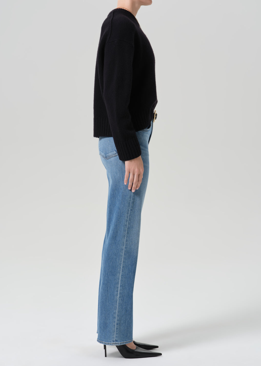 Person wearing a black crewneck sweater with light wash straight leg jeans viewed from the side on a grey background
