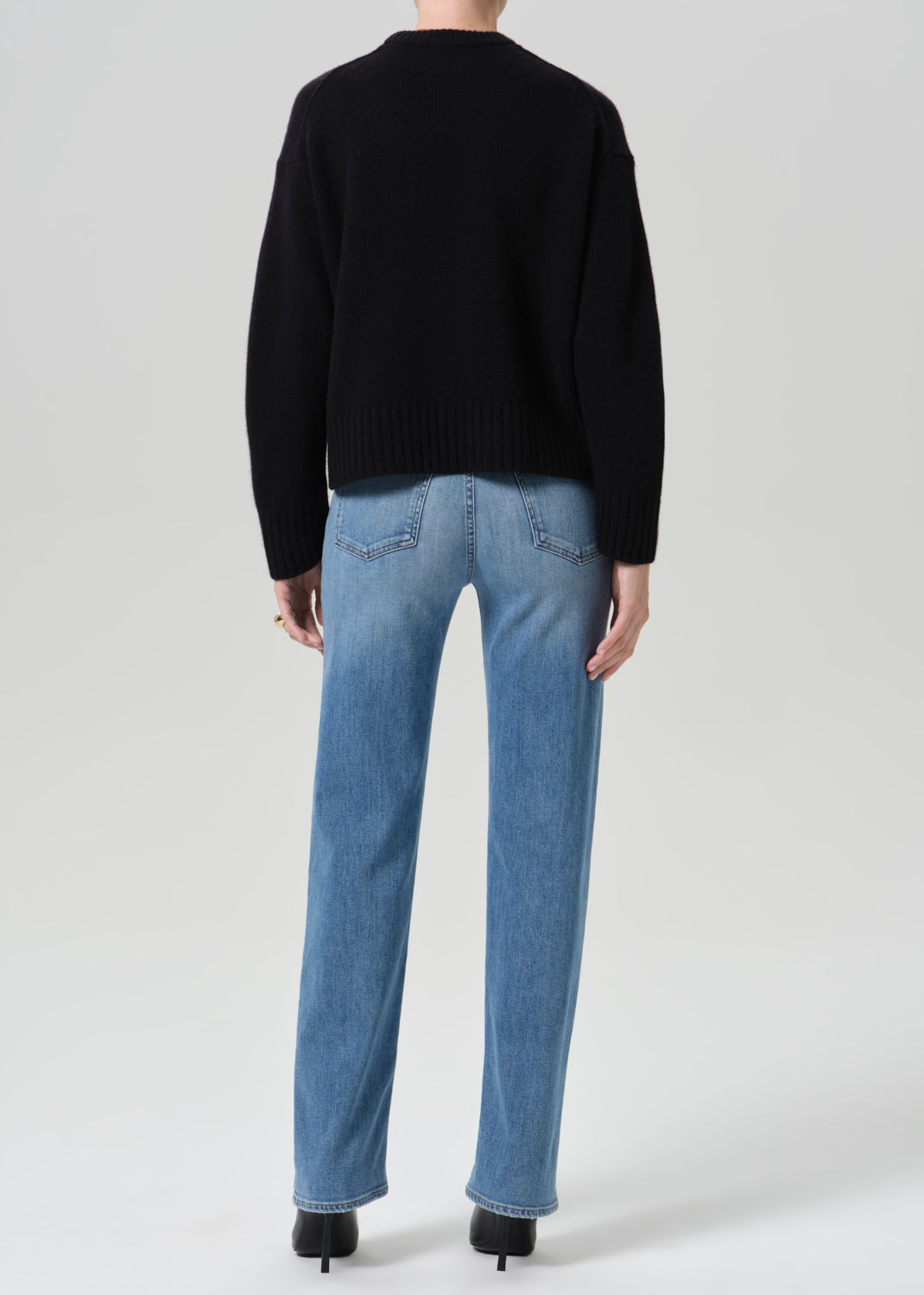 Person wearing a black crewneck sweater with light wash straight leg jeans viewed from the back on a grey background