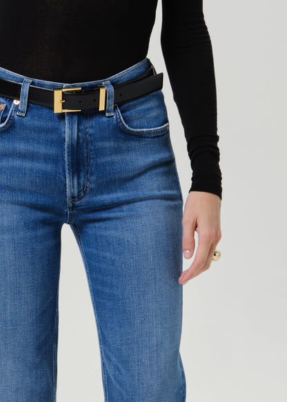 A close-up of a person wearing blue jeans with a black leather belt featuring a gold-tone rectangular buckle, paired with a black long-sleeve top against a light gray studio background.