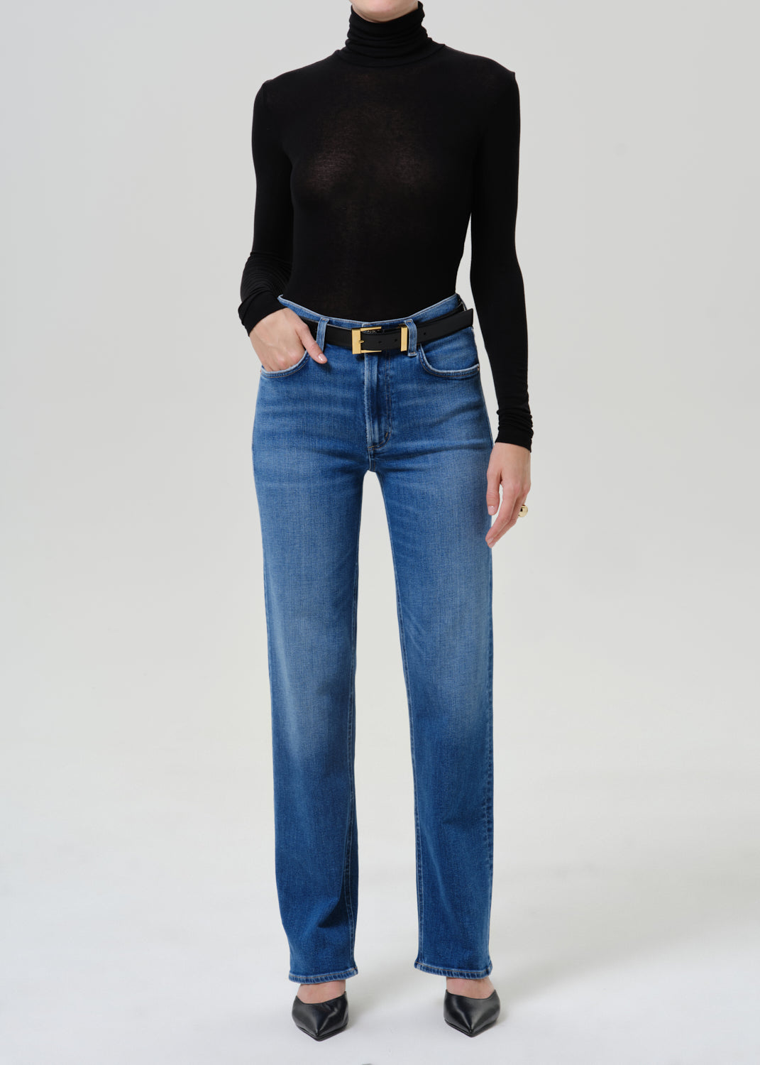 A model wearing a black long-sleeve turtleneck top, blue straight-leg jeans with a black belt featuring a gold buckle, and black pointed-toe heels, standing against a light gray studio background.