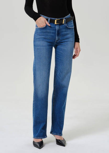 A model wearing a pair of high-waisted, straight-leg blue jeans with a black leather belt featuring a gold buckle, styled with a black long-sleeve top and black pointed-toe heels against a light gray studio background.