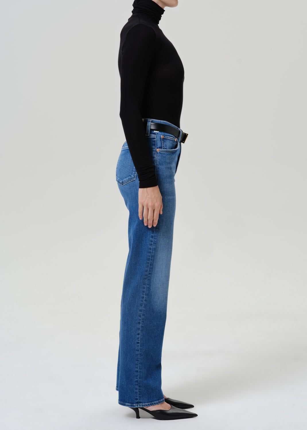 A side-profile studio shot of a model wearing high-waisted, wide-leg blue jeans, a black long-sleeve turtleneck, and black pointed-toe heels against a light gray background.