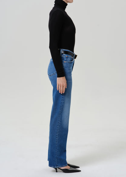 A side-profile studio shot of a model wearing high-waisted, wide-leg blue jeans, a black long-sleeve turtleneck, and black pointed-toe heels against a light gray background.