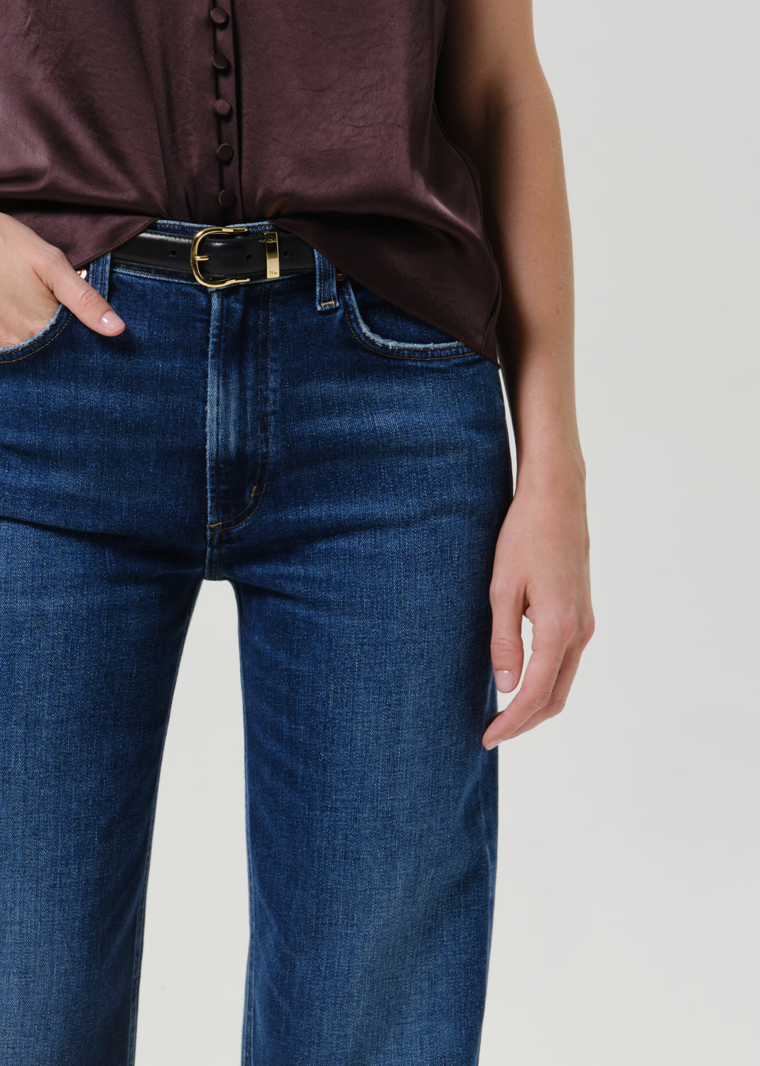 Close-up of a person wearing dark blue denim jeans with a black belt featuring a gold buckle, paired with a dark brown satin top, against a light gray studio background.