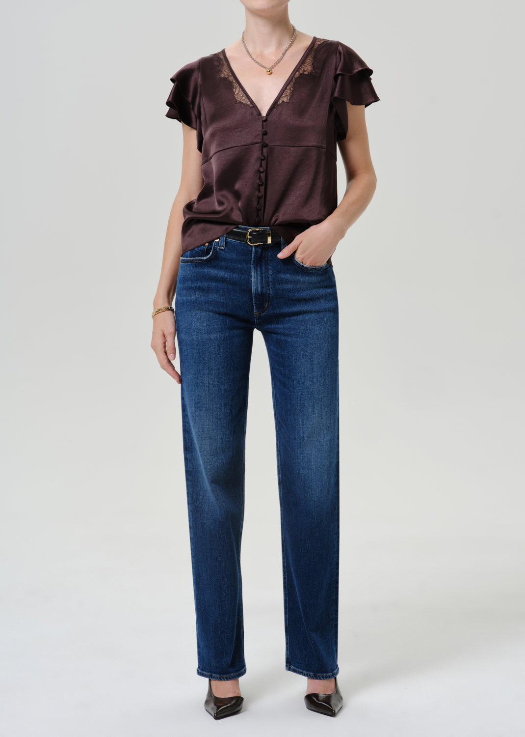 A model wearing a dark brown ruffled short-sleeve blouse with lace trim and dark blue straight-leg jeans, standing against a light gray studio background.