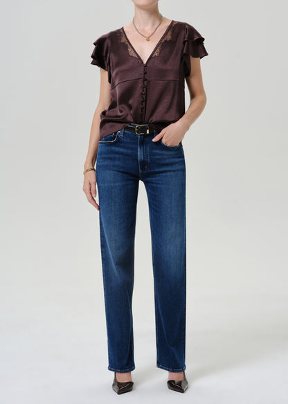 A model wearing a dark brown ruffled short-sleeve blouse with lace trim and dark blue straight-leg jeans, standing against a light gray studio background.