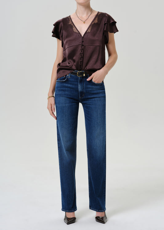 A model wearing a dark brown ruffled short-sleeve blouse with lace trim and dark blue straight-leg jeans, standing against a light gray studio background.