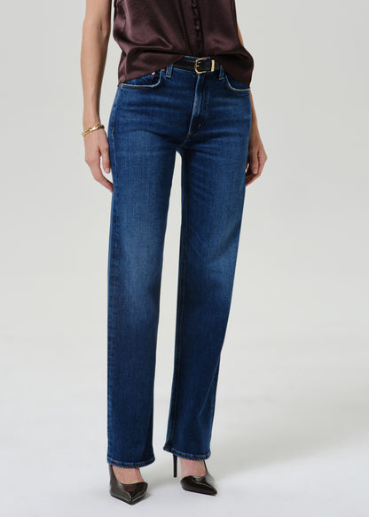 A model wearing dark blue straight-leg jeans with a brown satin blouse and black pointed-toe heels, standing against a light gray studio background.