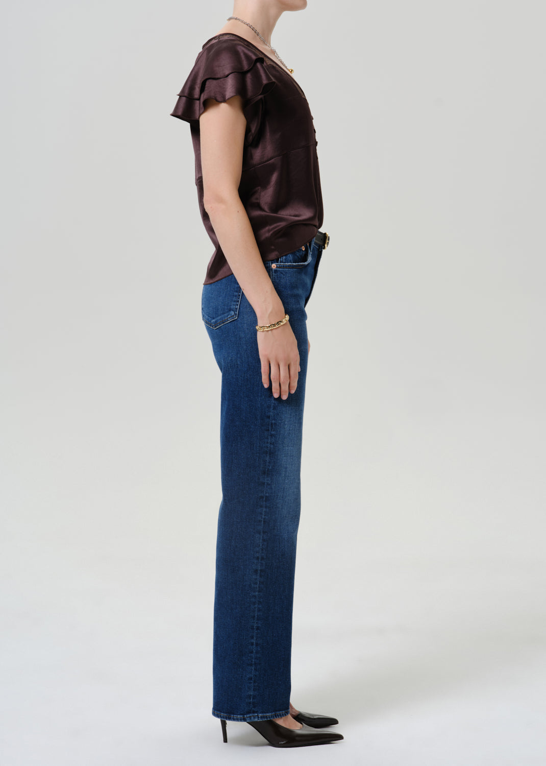 A side-profile studio shot of a model wearing a dark brown ruffled short-sleeve top, blue jeans, and black heels against a light gray background.