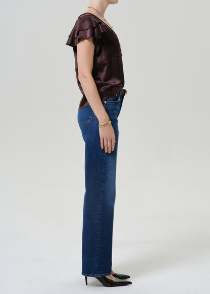 A side-profile studio shot of a model wearing a dark brown ruffled short-sleeve top, blue jeans, and black heels against a light gray background.