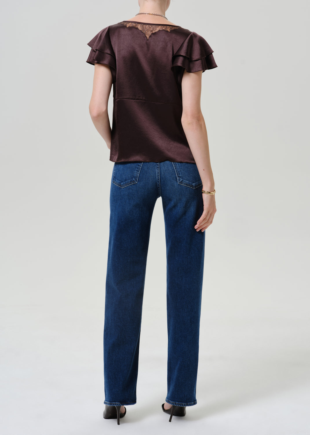 A model shown from the back wearing a dark brown ruffled short-sleeve top and blue jeans against a light gray studio background.