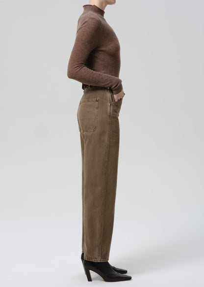 Person wearing a brown turtleneck and beige pants on a grey background