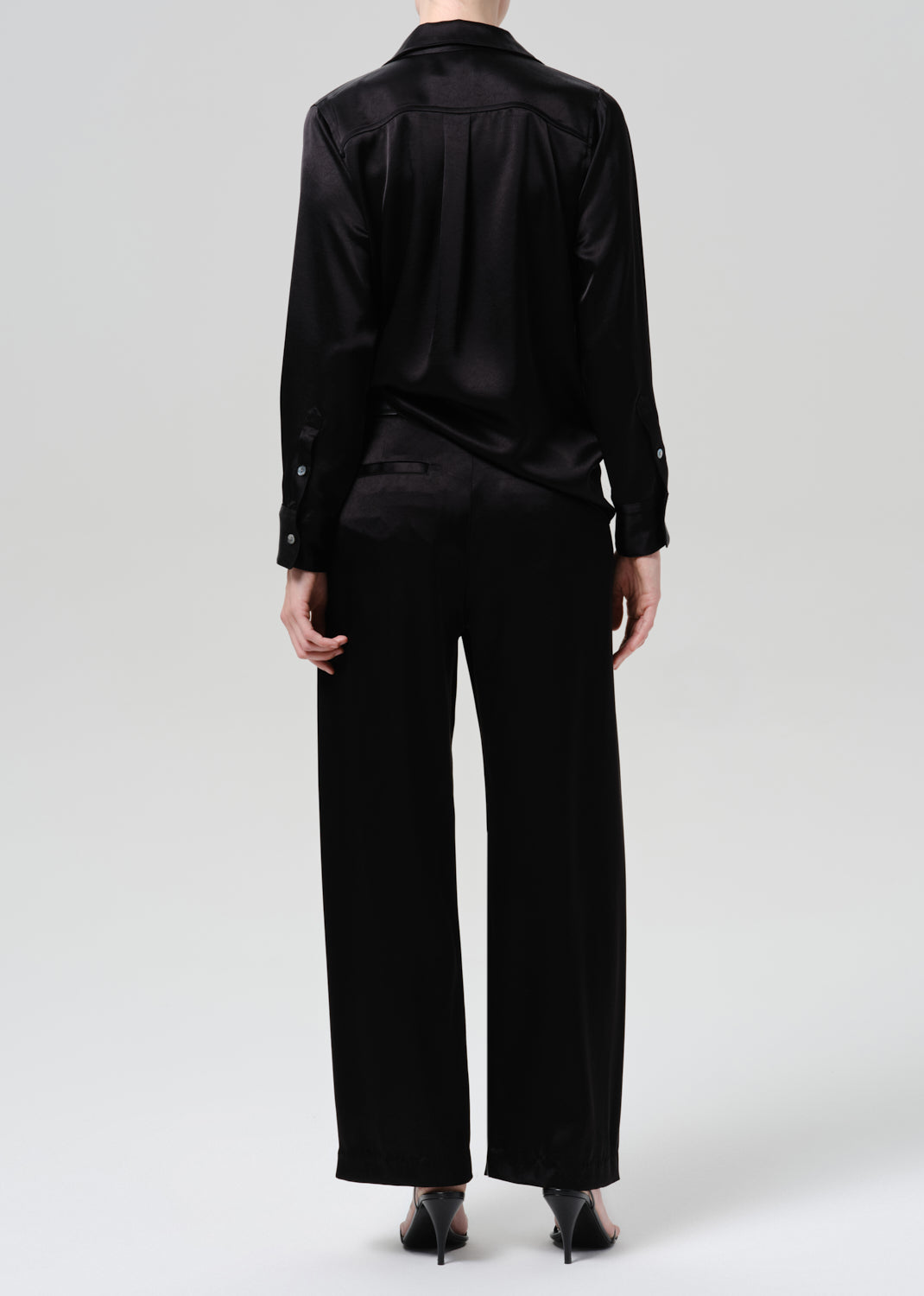 Person wearing a black satin button front blouse with black wide leg trousers viewed from the back on a grey background