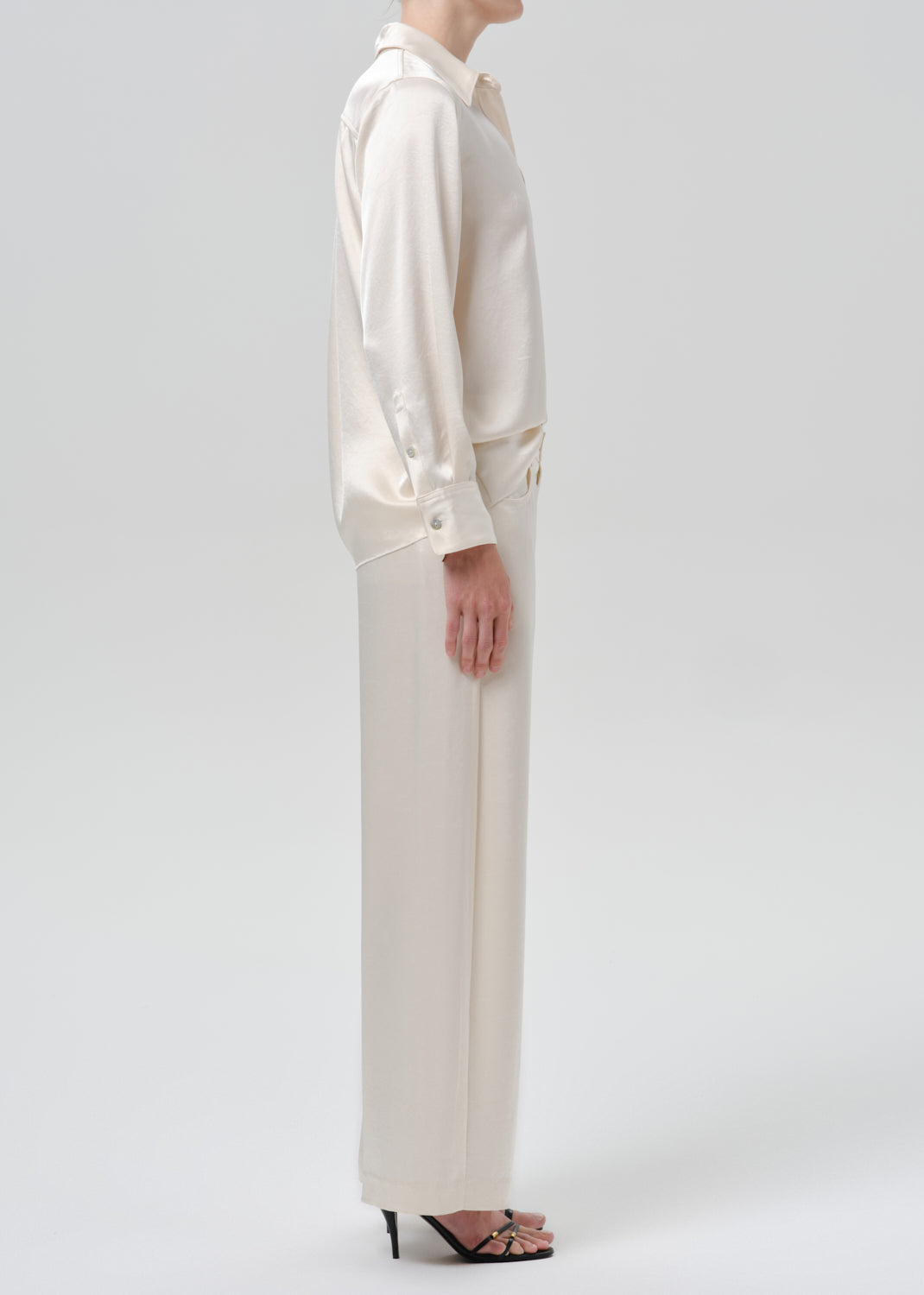 Person wearing an ivory satin button front blouse with ivory wide leg trousers viewed from the side on a grey background