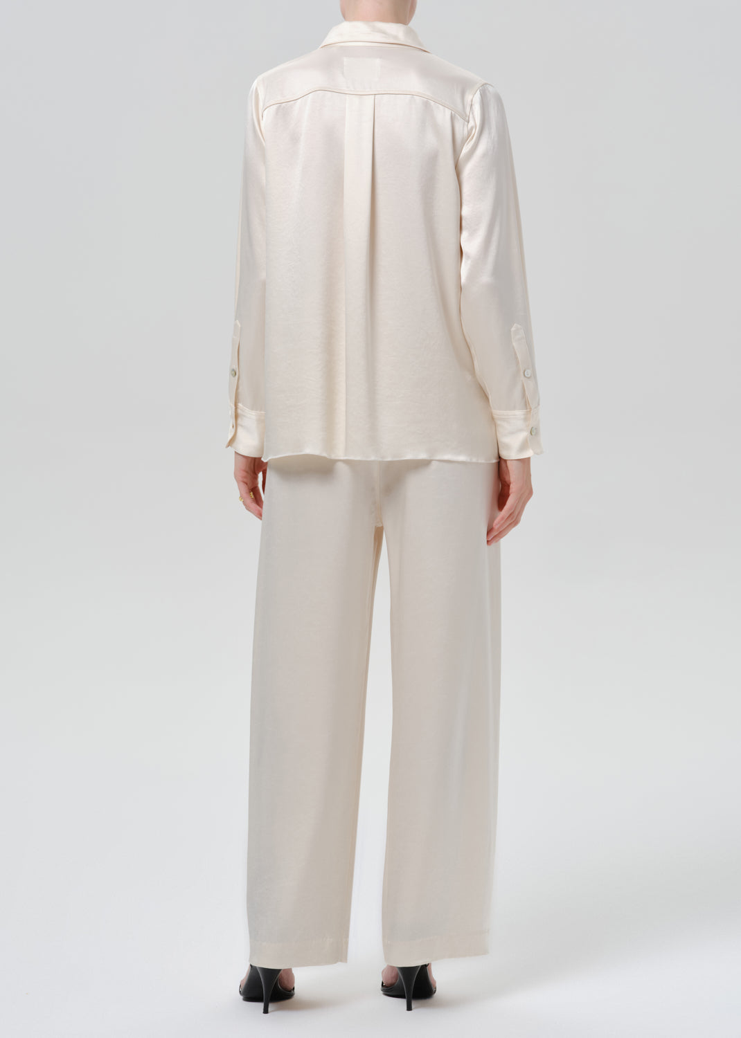 Person wearing an ivory satin button front blouse with ivory wide leg trousers viewed from the back on a grey background