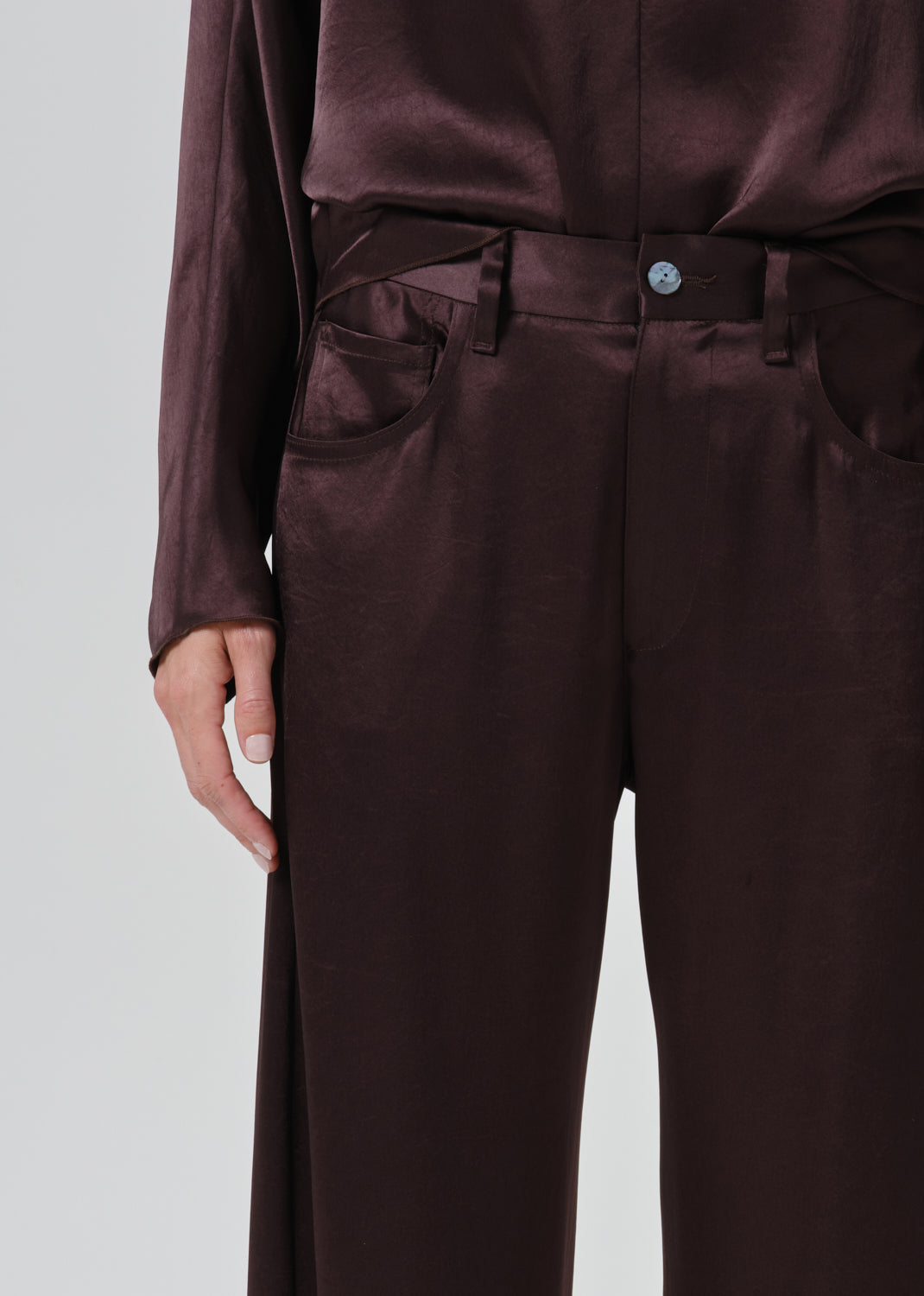 Close-up of a person wearing dark brown satin trousers with a matching top, showing the waist, belt, front pockets, and hand against a grey background.