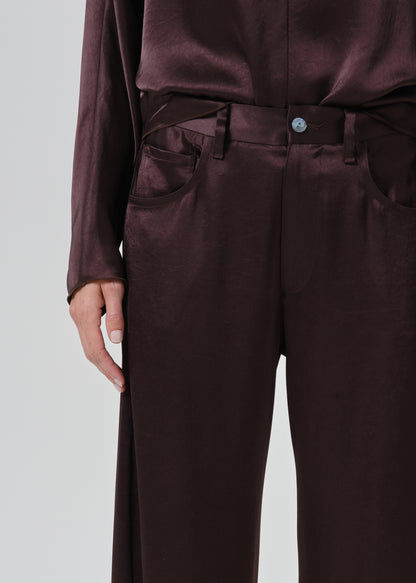 Close-up of a person wearing dark brown satin trousers with a matching top, showing the waist, belt, front pockets, and hand against a grey background.