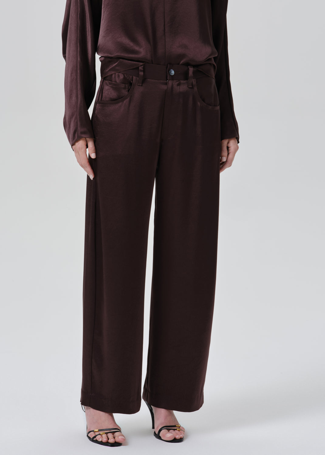A model wearing dark brown wide-leg trousers with a matching belt and waist tie, paired with a matching dark brown satin long-sleeve top and black strappy heels, against a grey background.