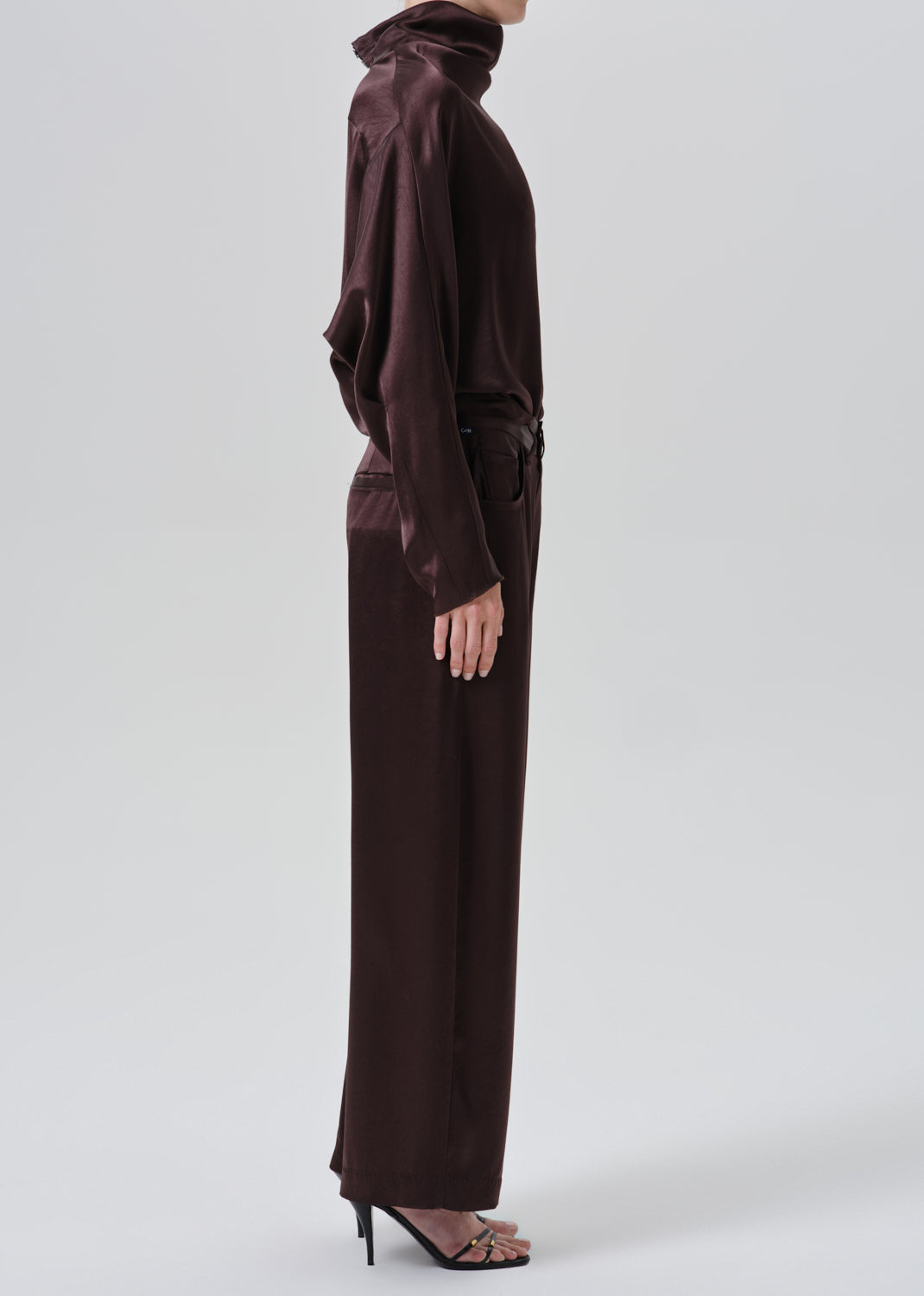 A model shown from the side wearing a dark brown satin turtleneck top and matching wide-leg trousers with a tie belt at the waist, paired with black high-heeled sandals, against a grey background.