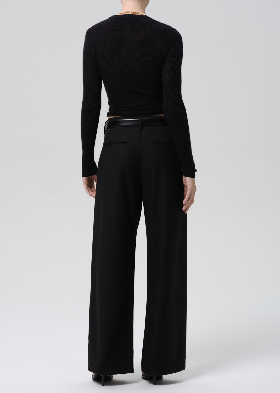 Person wearing a black fitted long sleeve sweater with black wide leg trousers viewed from the back on a grey background