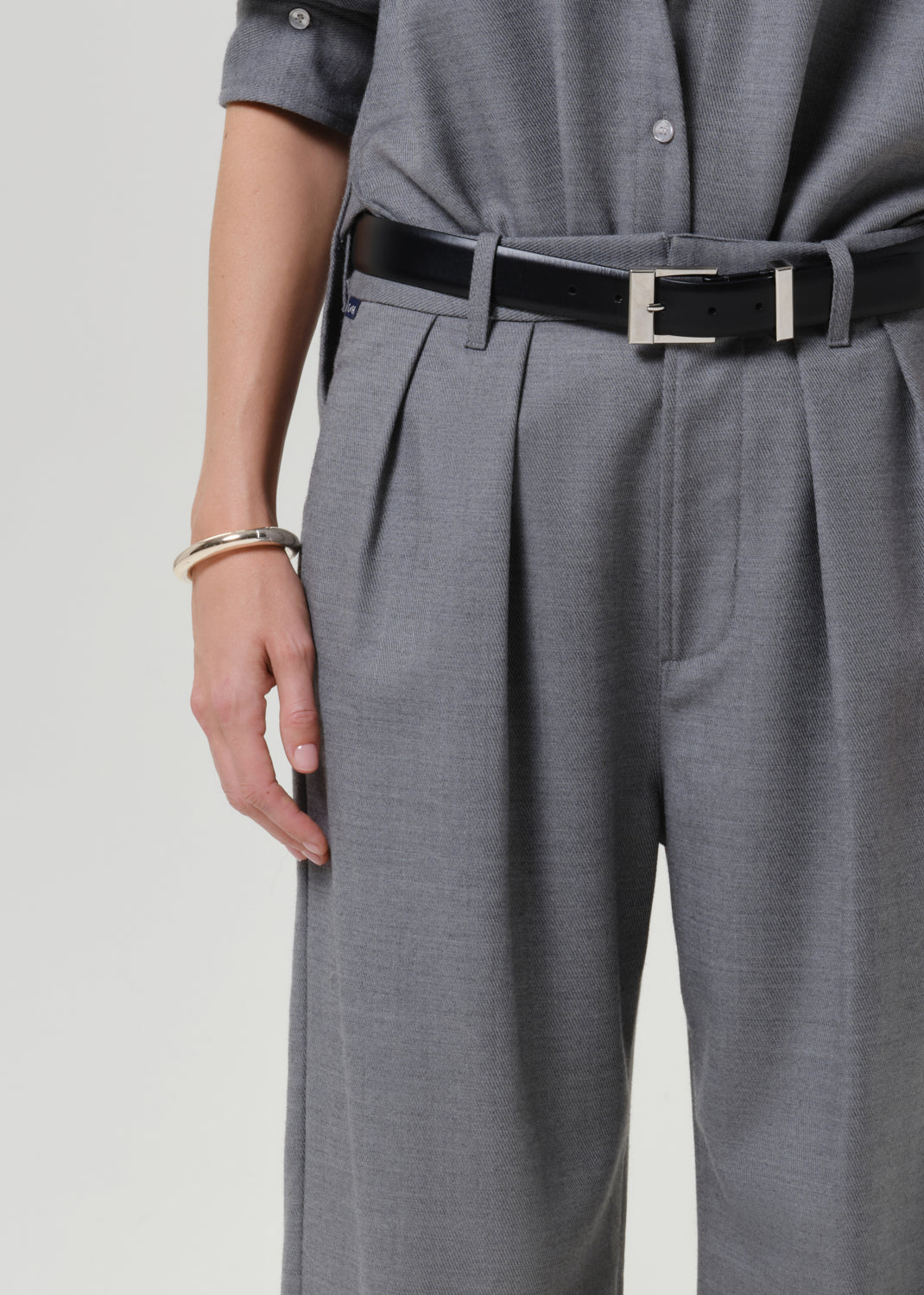 Close-up of a person wearing gray pleated trousers with a black belt featuring a silver rectangular buckle, paired with a matching gray top.