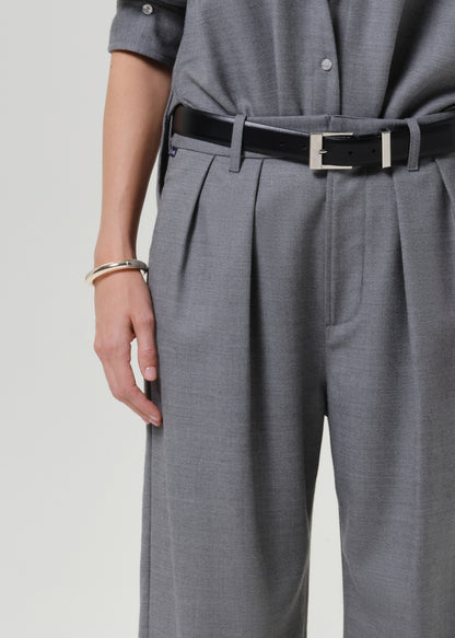 Close-up of a person wearing gray pleated trousers with a black belt featuring a silver rectangular buckle, paired with a matching gray top.