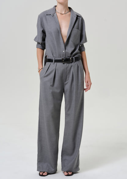 A model wearing a gray short-sleeve collared jumpsuit with wide-leg trousers, pleated details, and a black belt, photographed against a light gray studio background.