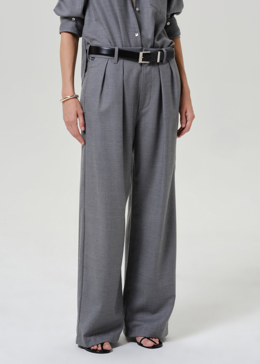 A model wearing high-waisted, wide-leg gray trousers with pleats, styled with a black leather belt, a gray shirt, and black sandals against a light gray studio background.