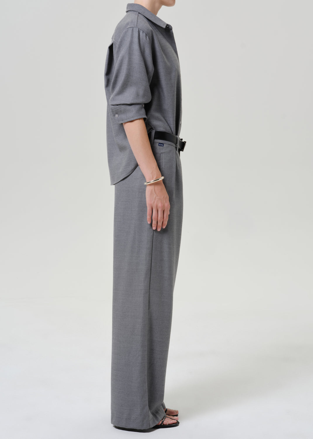A model shown from the side wearing a gray long-sleeve collared shirt and matching wide-leg trousers, styled with a black belt and black sandals against a light gray studio background.