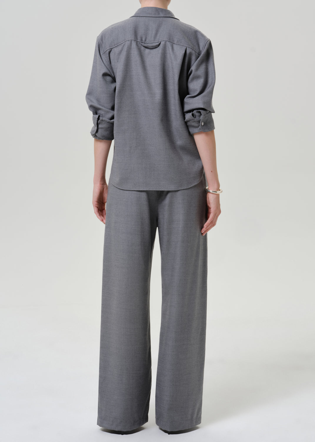 A model shown from the back wearing a gray two-piece set consisting of a long-sleeve button-down shirt and matching wide-leg trousers, photographed against a neutral light gray studio background.