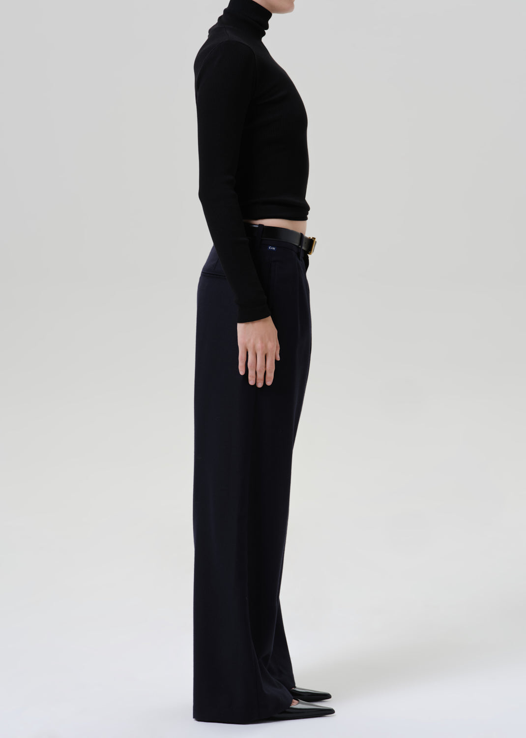 Person wearing a black fitted turtleneck with dark navy wide leg trousers viewed from the side on a grey background