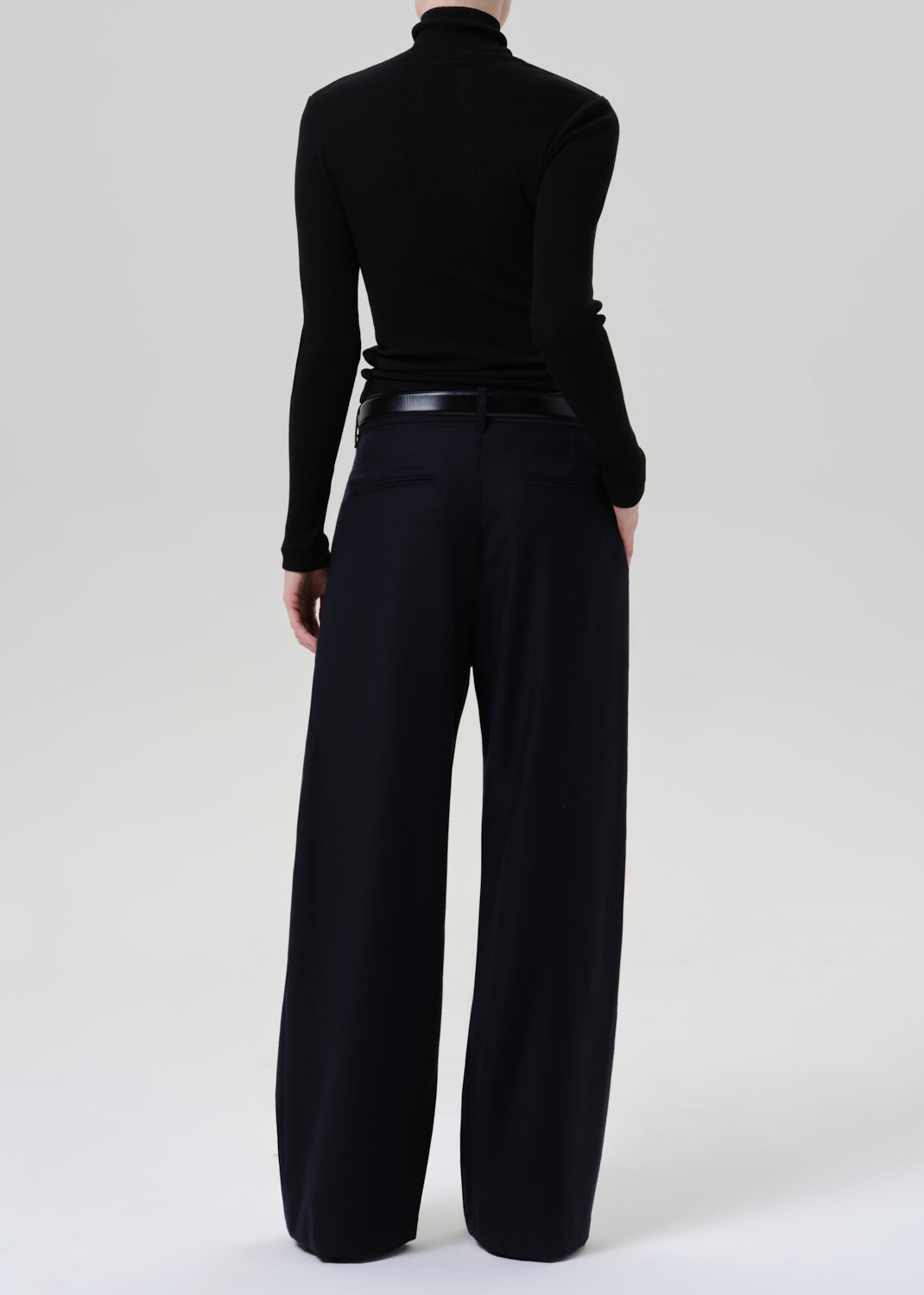 Person wearing a black fitted turtleneck with dark navy wide leg trousers viewed from the back on a grey background