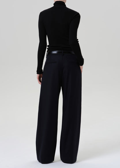 Person wearing a black fitted turtleneck with dark navy wide leg trousers viewed from the back on a grey background