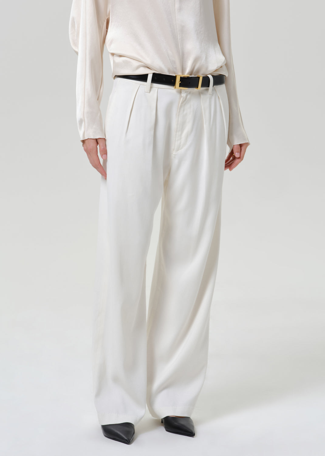 Woman wearing white wide leg pants with an ivory satin draped neck top on a grey background.