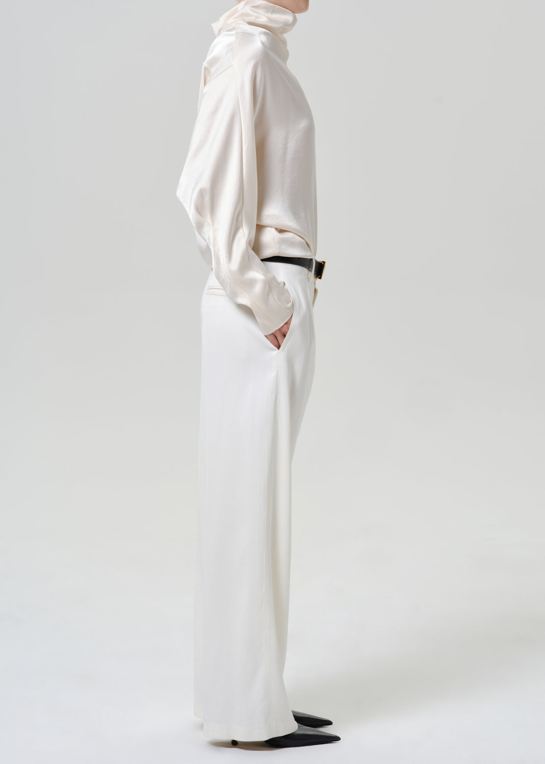 Woman wearing white wide leg pants with an ivory satin draped neck top viewed from the side on a grey background.