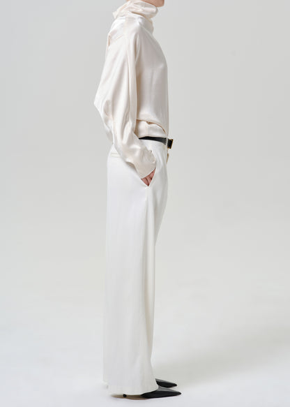 Woman wearing white wide leg pants with an ivory satin draped neck top viewed from the side on a grey background.