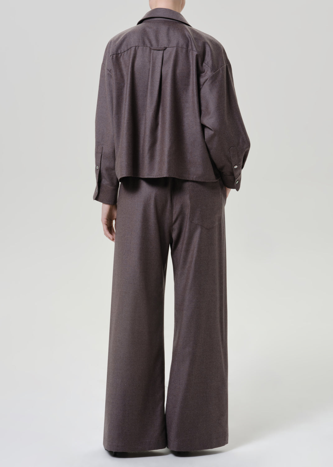 Woman wearing dark brown wide leg pants with a matching dark brown short sleeve top viewed from the back on a grey background.