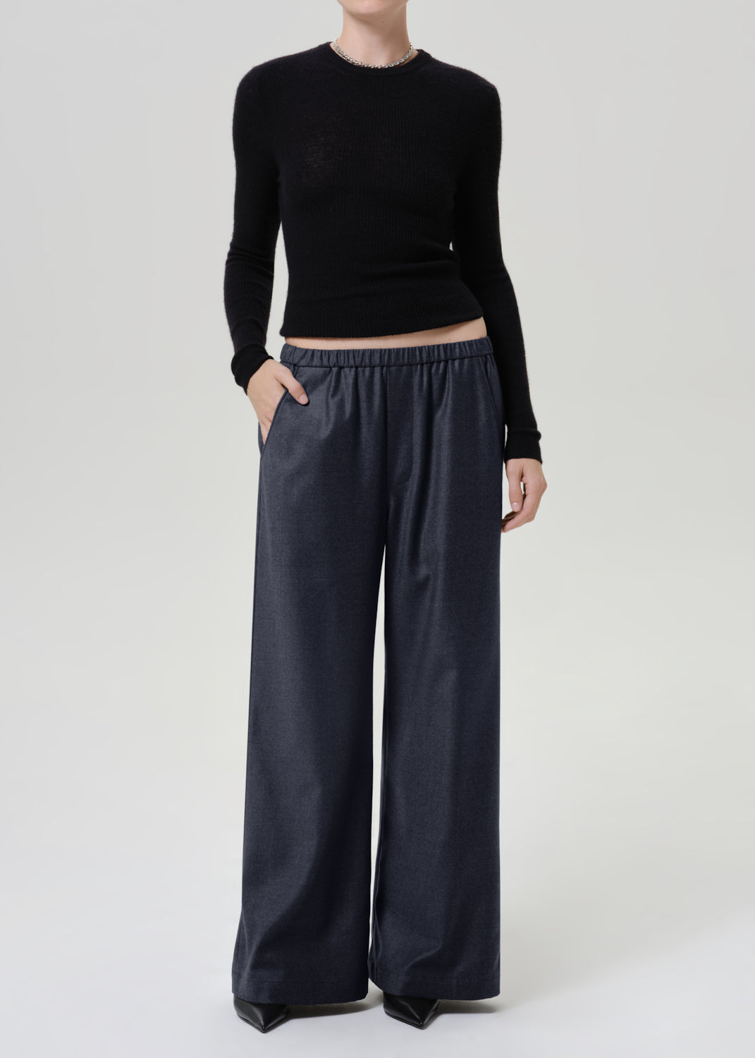 Woman wearing dark grey wide leg pants with a black long sleeve top on a grey background.