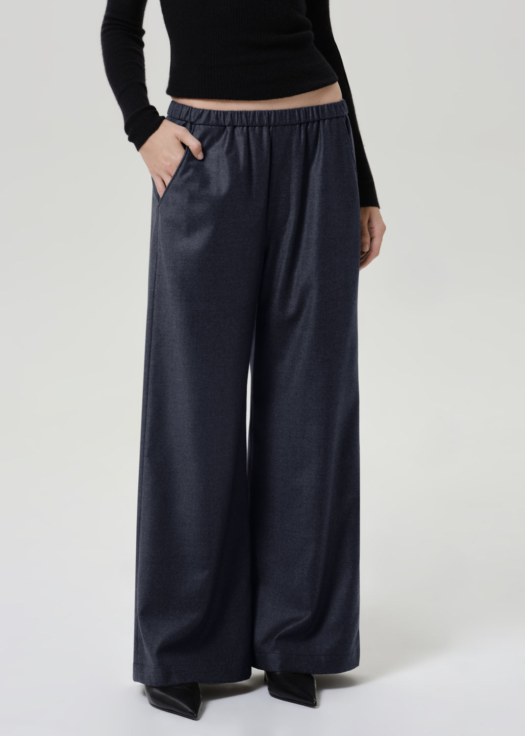 Woman wearing dark grey wide leg pants with a black long sleeve top on a grey background.
