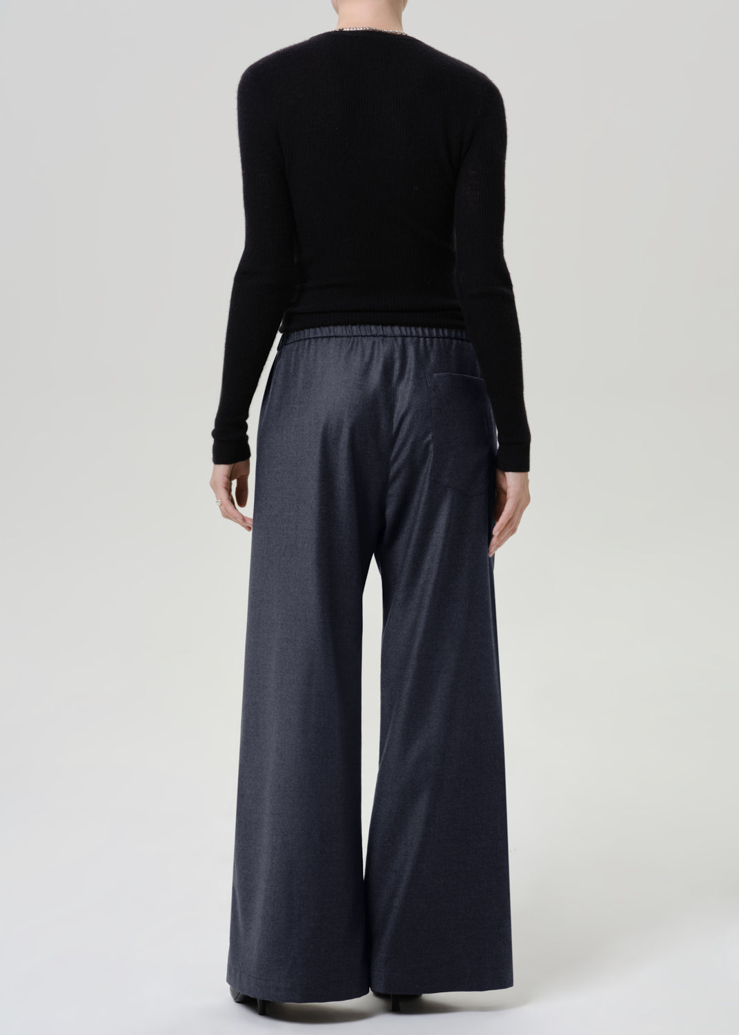 Woman wearing dark grey wide leg pants with a black long sleeve top viewed from the back on a grey background.