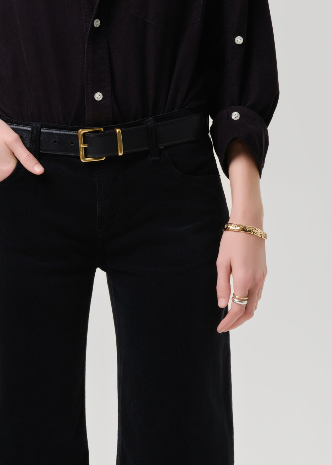Person wearing a black outfit with a belt and gold accessories on a grey background