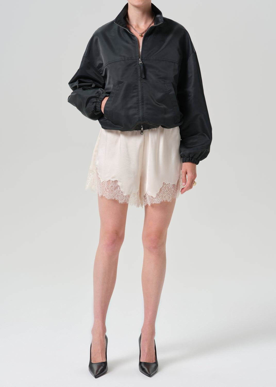 A model wearing a black zip-up bomber jacket, cream lace-trimmed shorts, and black pointed-toe heels, photographed against a grey background.